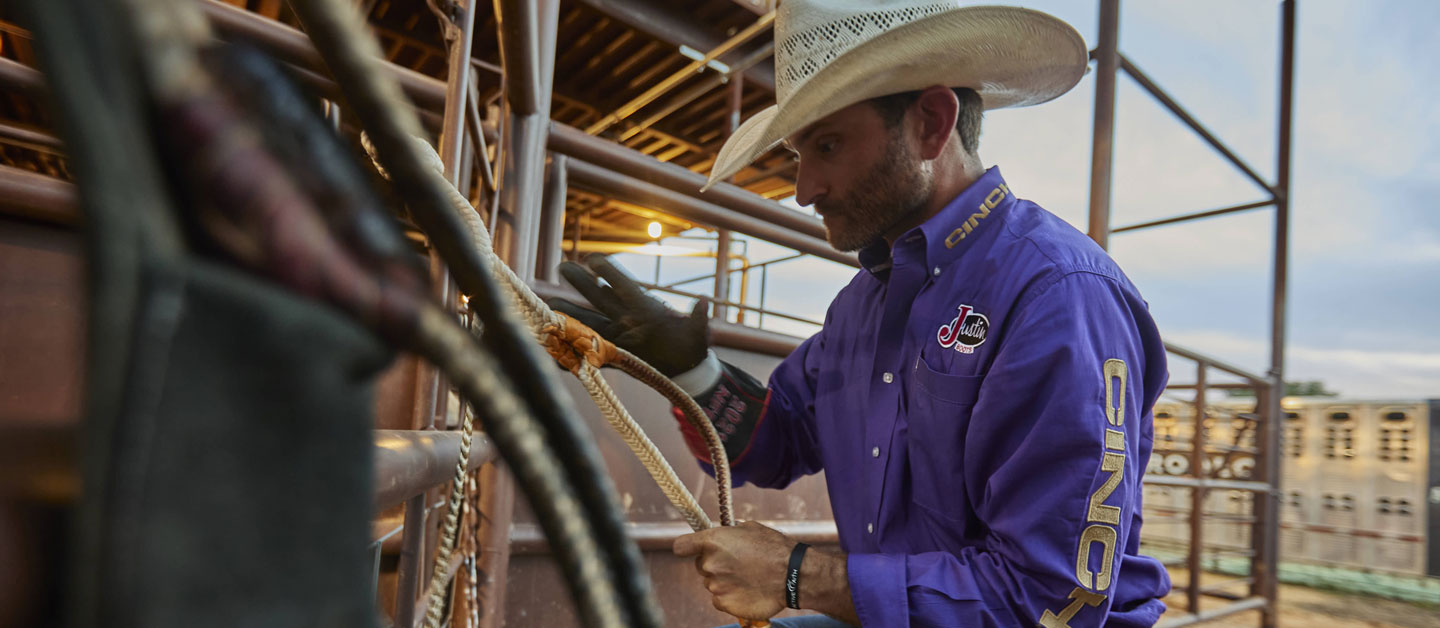 Man wearing a purple shirt, cowboy hat, and a glove getting ready to ride a bull by warming up the Rossin on his bull rope.